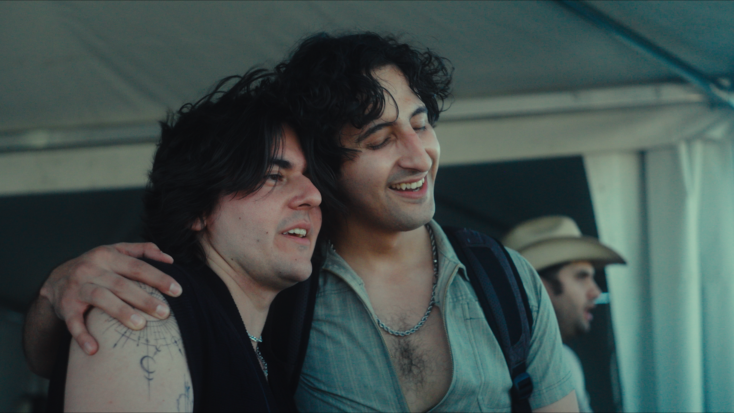 Two men with dark, curly hair sharing a moment of happiness, smiling and embracing each other, with a third person in the background.