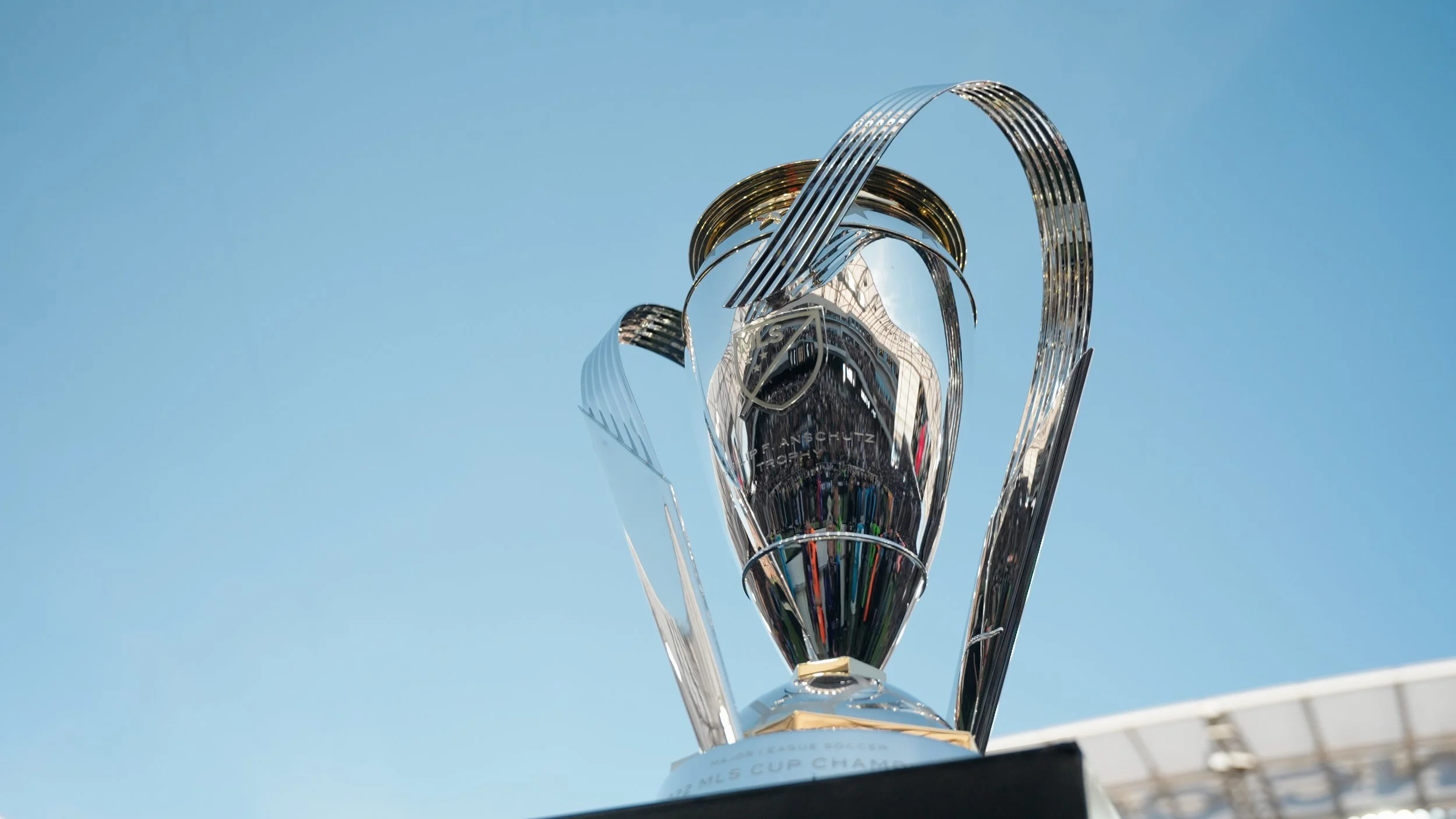 A shiny, metallic trophy cup with curved handles, standing on a white base against a clear blue sky.