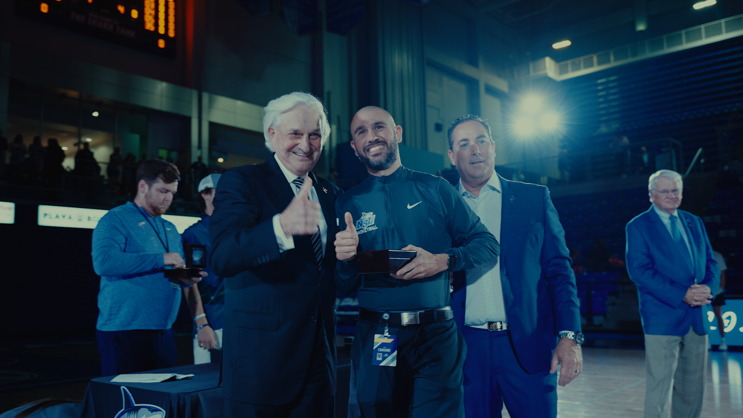 Group of men at an awards ceremony in an indoor sports arena, with one man receiving a medal, surrounded by officials and staff.