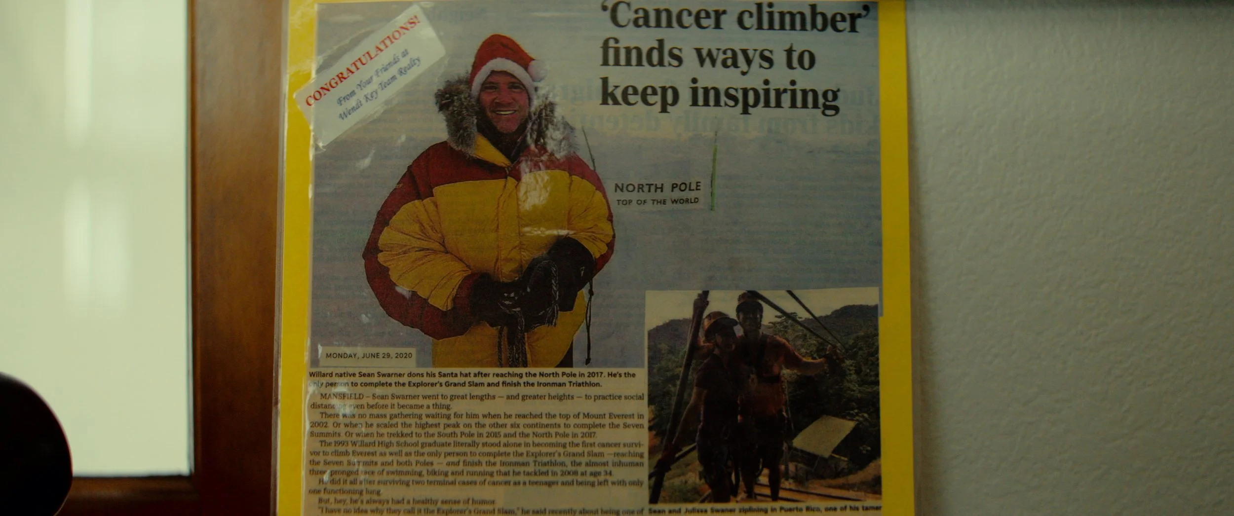 A newspaper clipping about a cancer survivor who reached the North Pole, featuring a man in a yellow and red winter jacket, wearing a Santa hat and smiling, with a smaller photo of him in a harness in a snowy landscape.