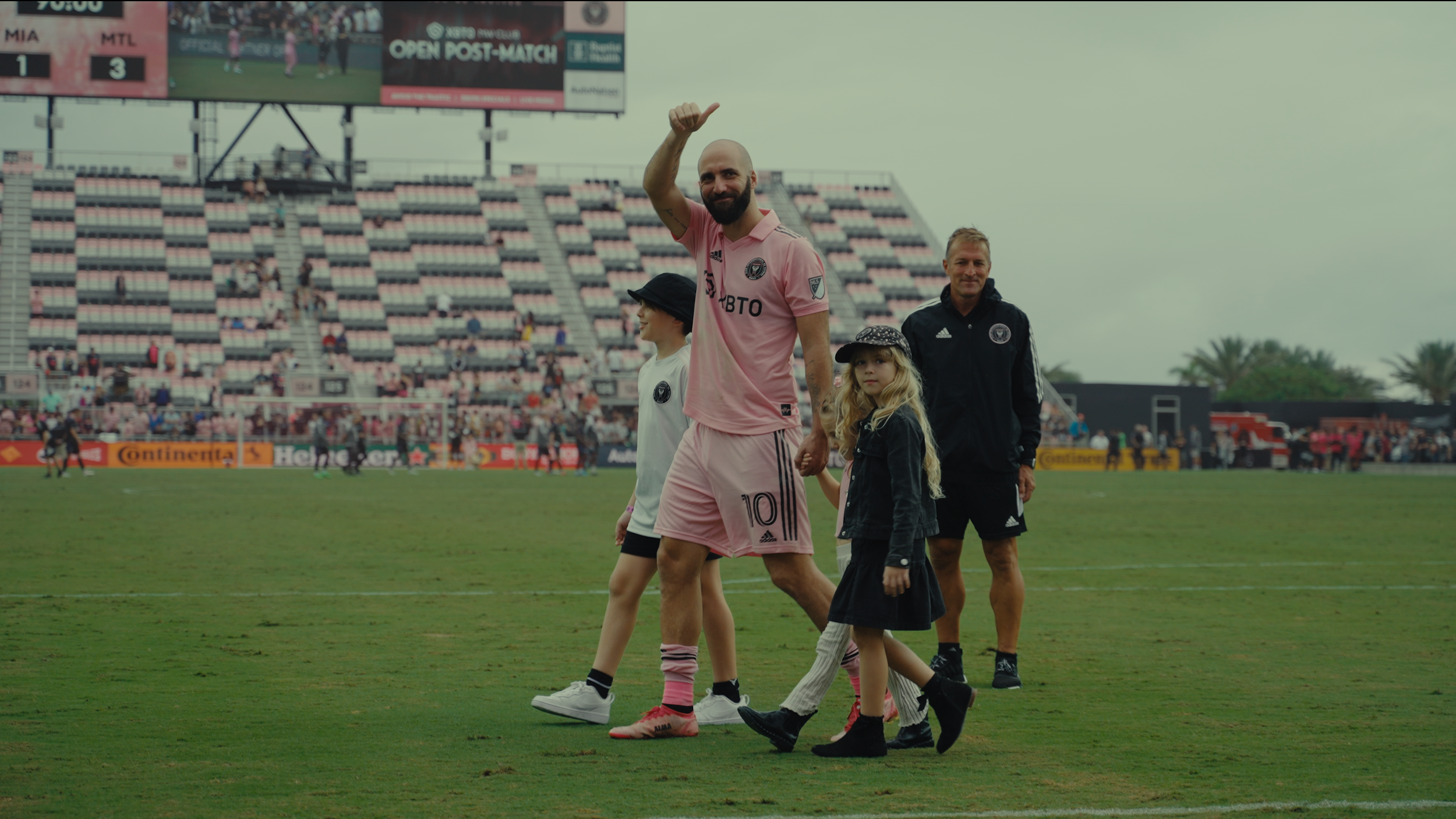 A soccer player in pink uniform waving to the crowd, holding hands with two children, walking on a soccer field with spectators in the background.