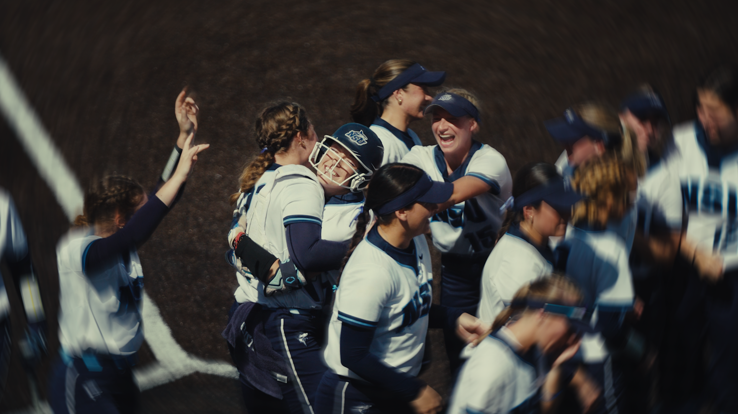 A group of female softball players celebrating on the field, smiling and hugging each other.