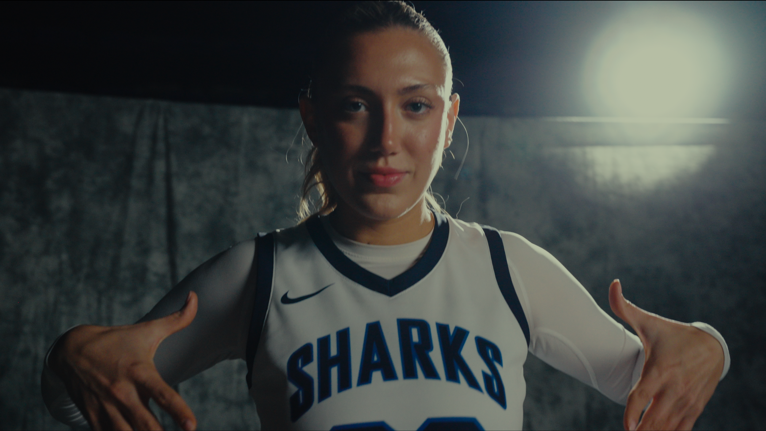 Female basketball player wearing a white jersey with the word 'SHARKS' on it, posing with hands near her hips, in front of dark background with bright studio lighting.