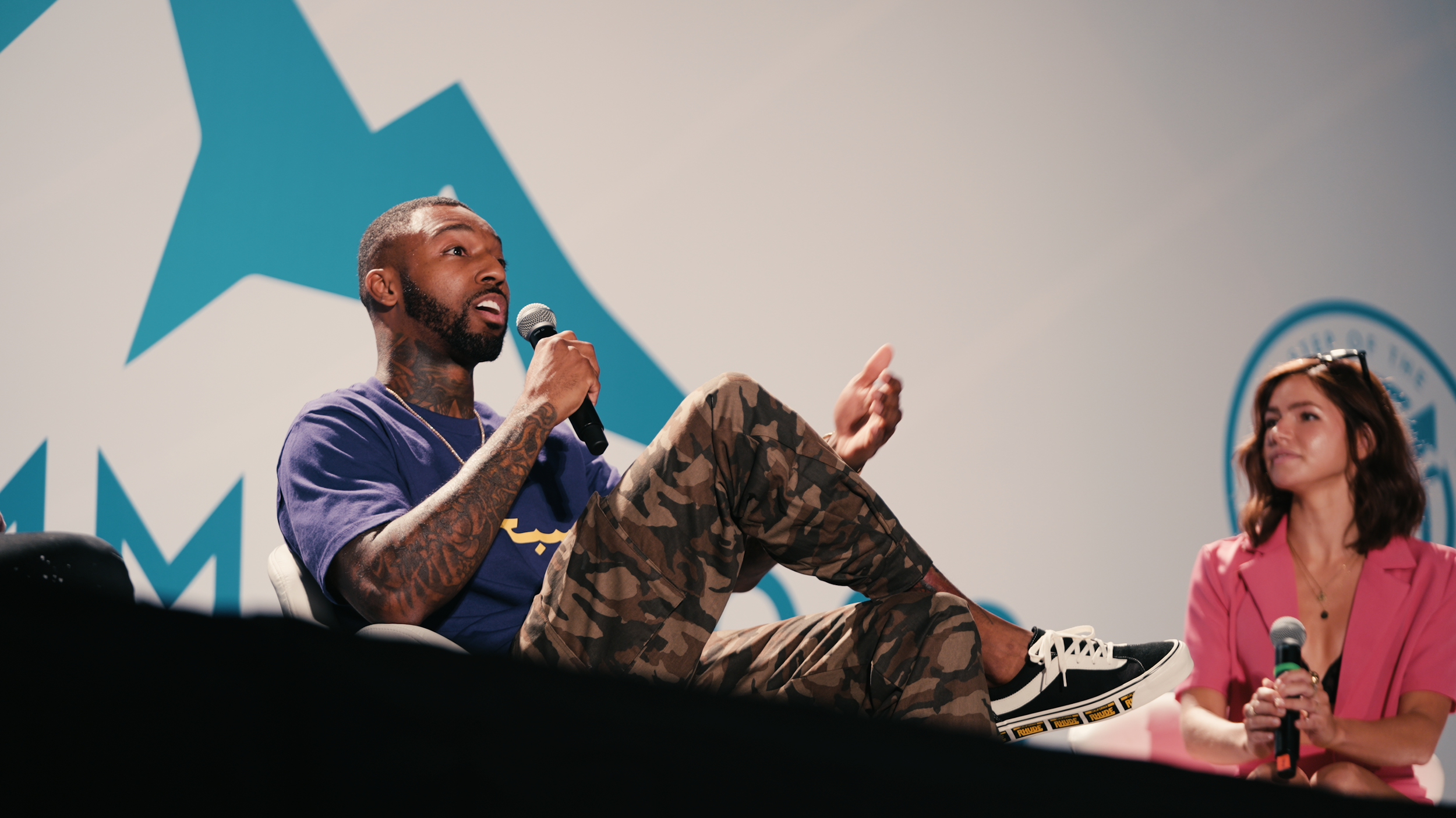 A man and woman sitting on stage during a panel discussion. The man is speaking into a microphone, wearing a purple shirt, camouflage pants, and sneakers. The woman is listening, wearing a pink blazer and holding a microphone.