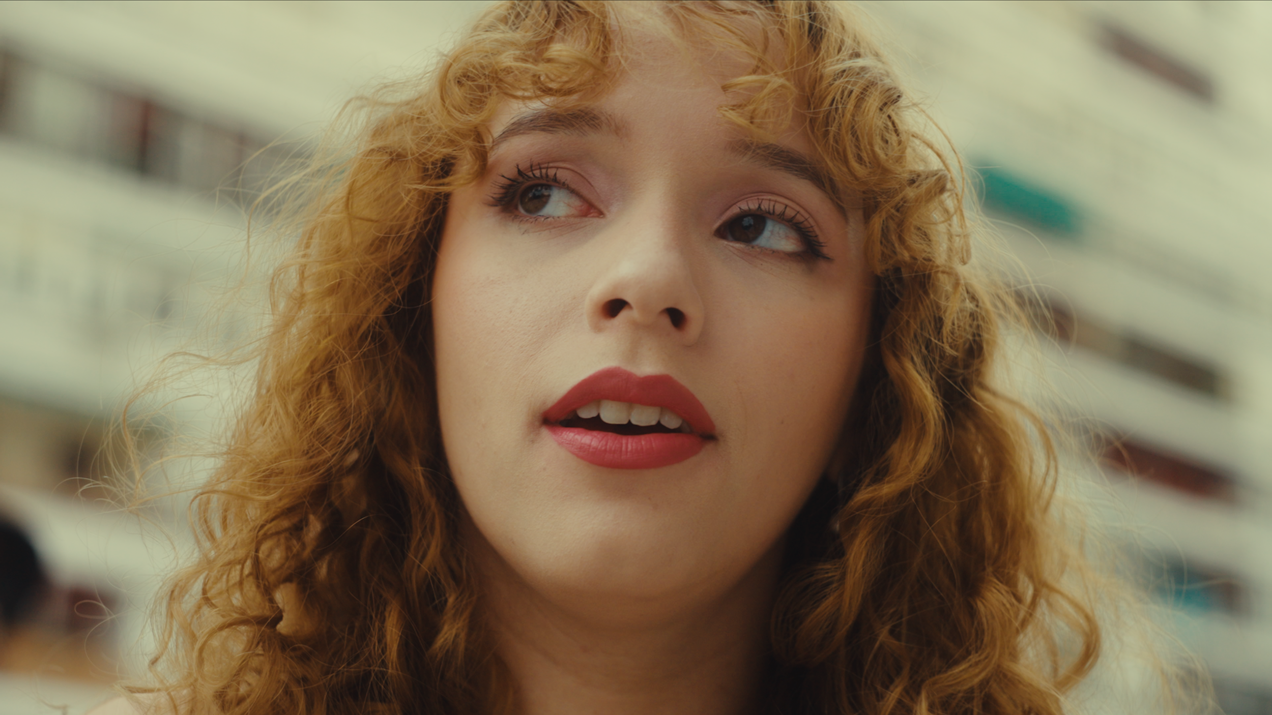 Close-up of a young woman with curly red hair, wearing makeup, looking to the side with an urban background.