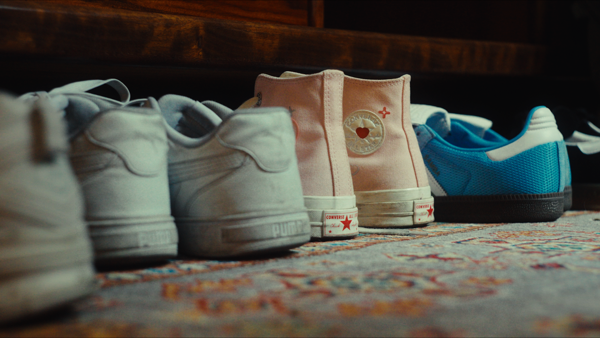 A row of sneakers and casual shoes lined up on a colorful rug, with a wooden piece of furniture in the background.