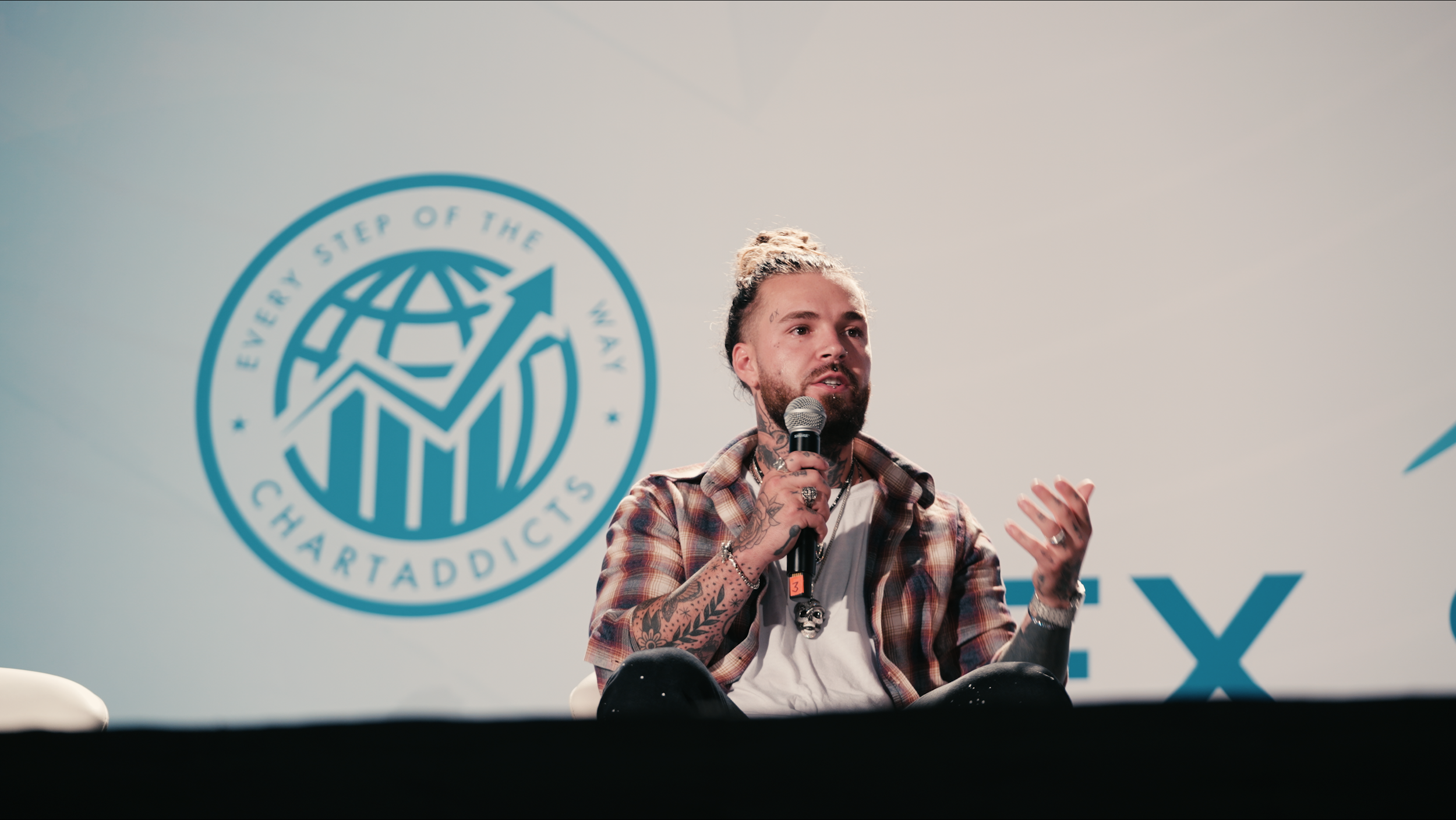 A man with tattoos on his arms and neck, wearing a plaid shirt and white t-shirt, is sitting on a stage holding a microphone. Behind him is a logo with a globe and an upward arrow, and the words 'Every Step of the Way' and 'ChartAddicts'.