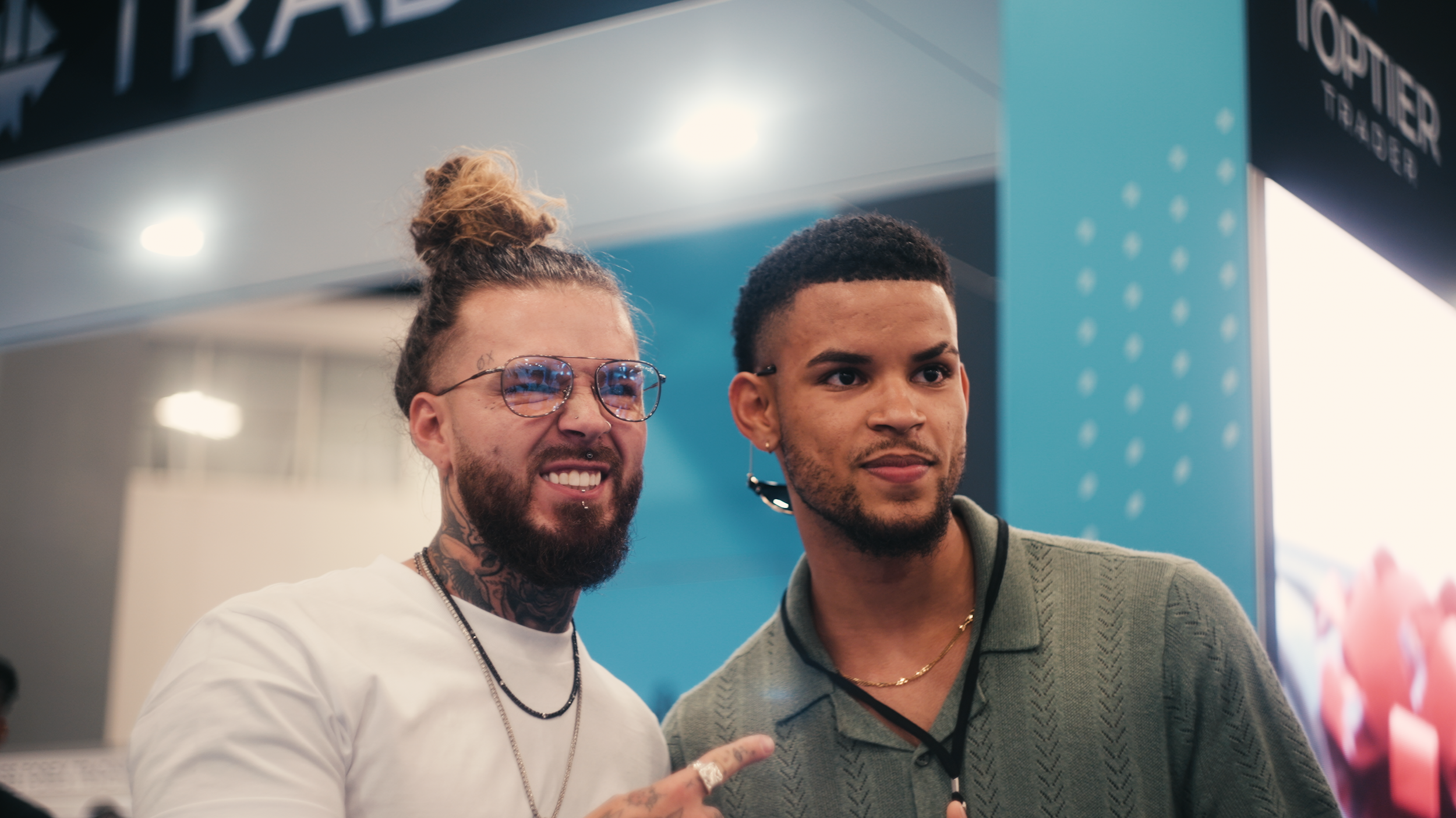 Two men posing together at an event, with one showing a tattoo on his neck, wearing glasses and a white shirt, and the other in a green sweater with a lanyard and earpiece. They are smiling and standing in front of a blue background with event signage.
