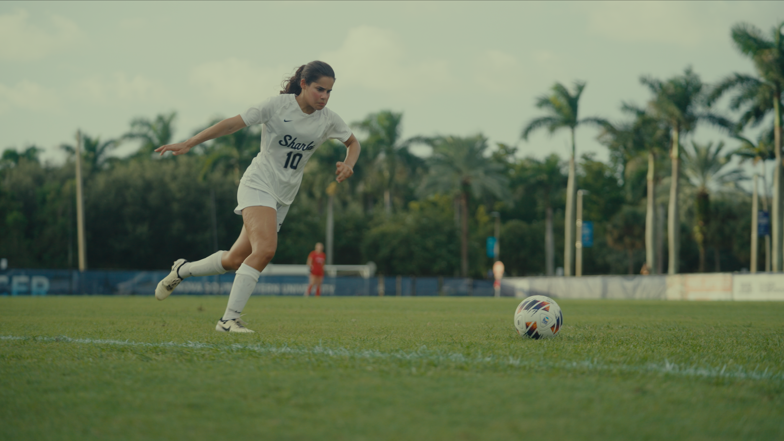A female soccer player wearing a white uniform with the number 10 on her jersey is running on a soccer field, about to kick a soccer ball. The background shows trees and a goalpost.