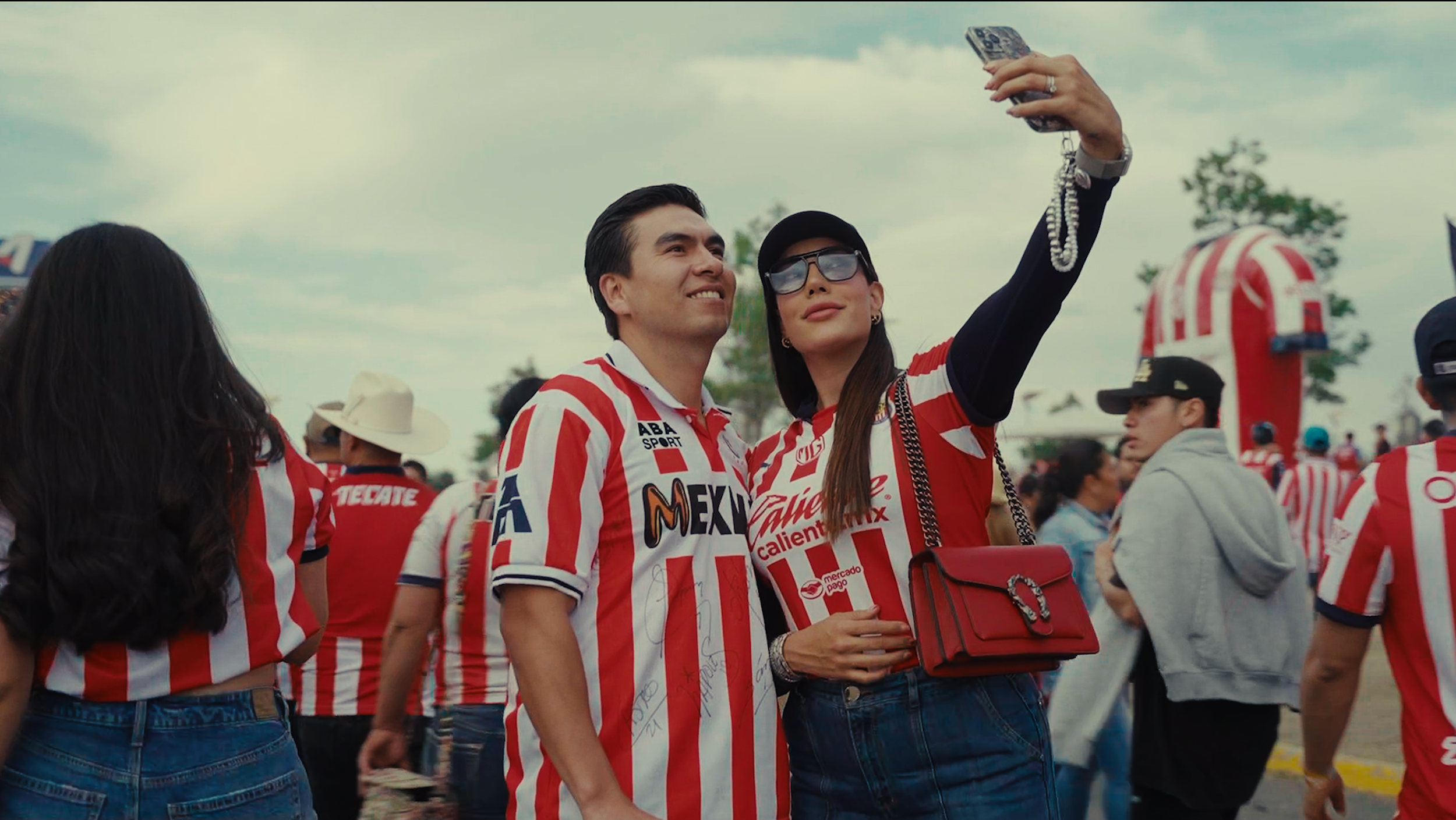 A man and woman wearing red and white striped soccer jerseys taking a selfie at a crowded outdoor event.