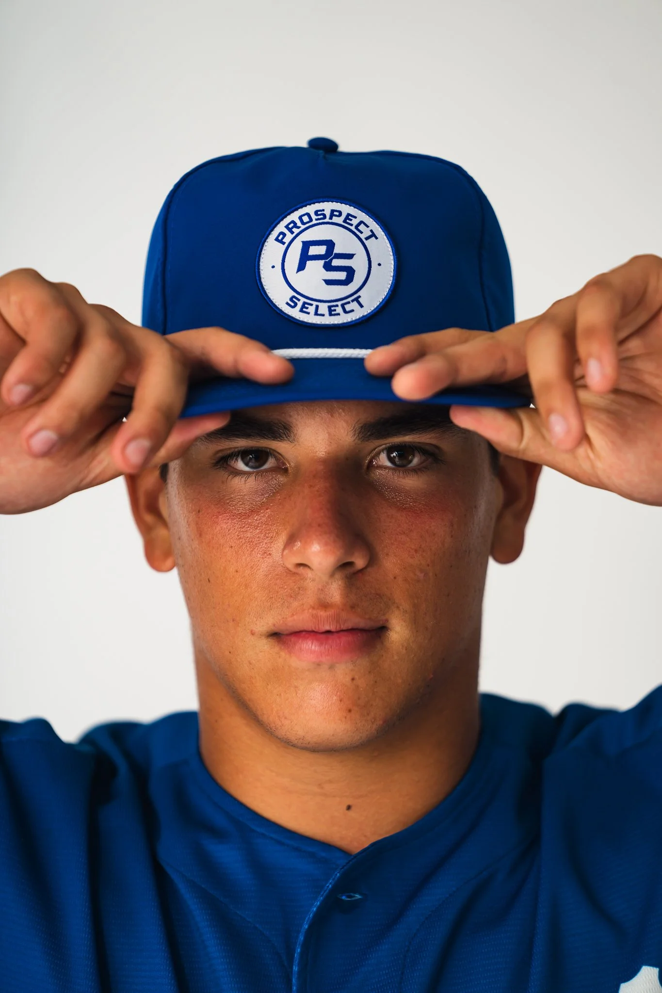 A young man wears a blue baseball cap with a logo that reads "Prospect Select" and "PS" on it. He is adjusting the cap with both hands and looking directly at the camera.