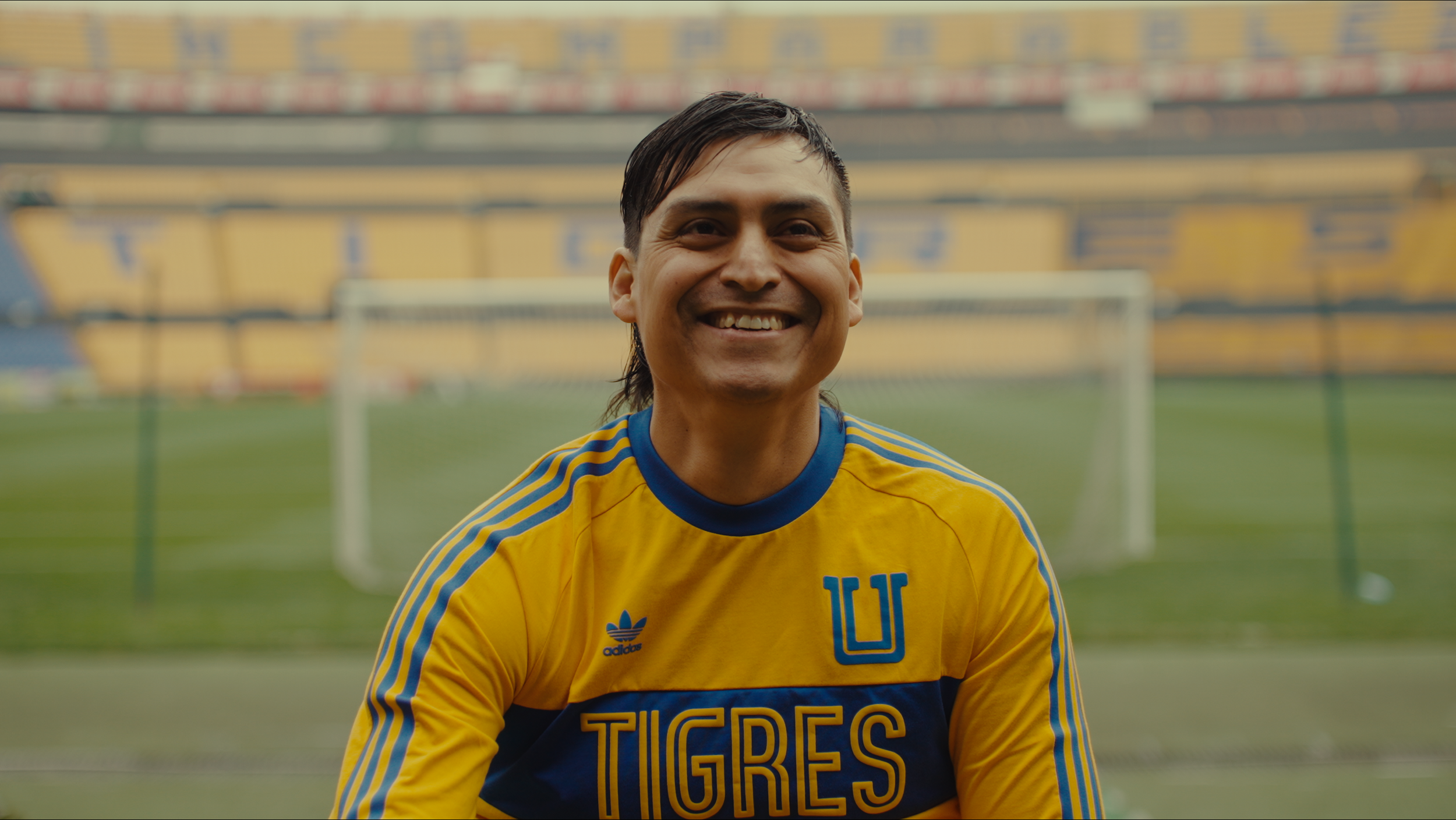 A smiling man wearing a yellow and blue soccer jersey with the word 'TIGRES' and a 'U' logo, standing in a soccer stadium with a goalpost in the background.
