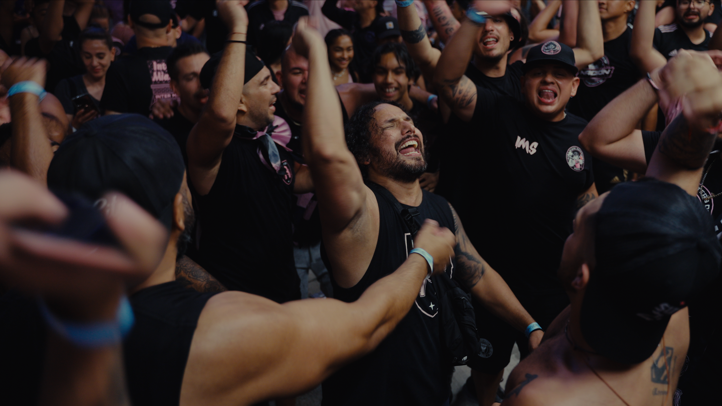 Crowd of diverse people at a concert or event, some with arms raised, cheering, and singing, dressed mostly in black, with excited expressions.