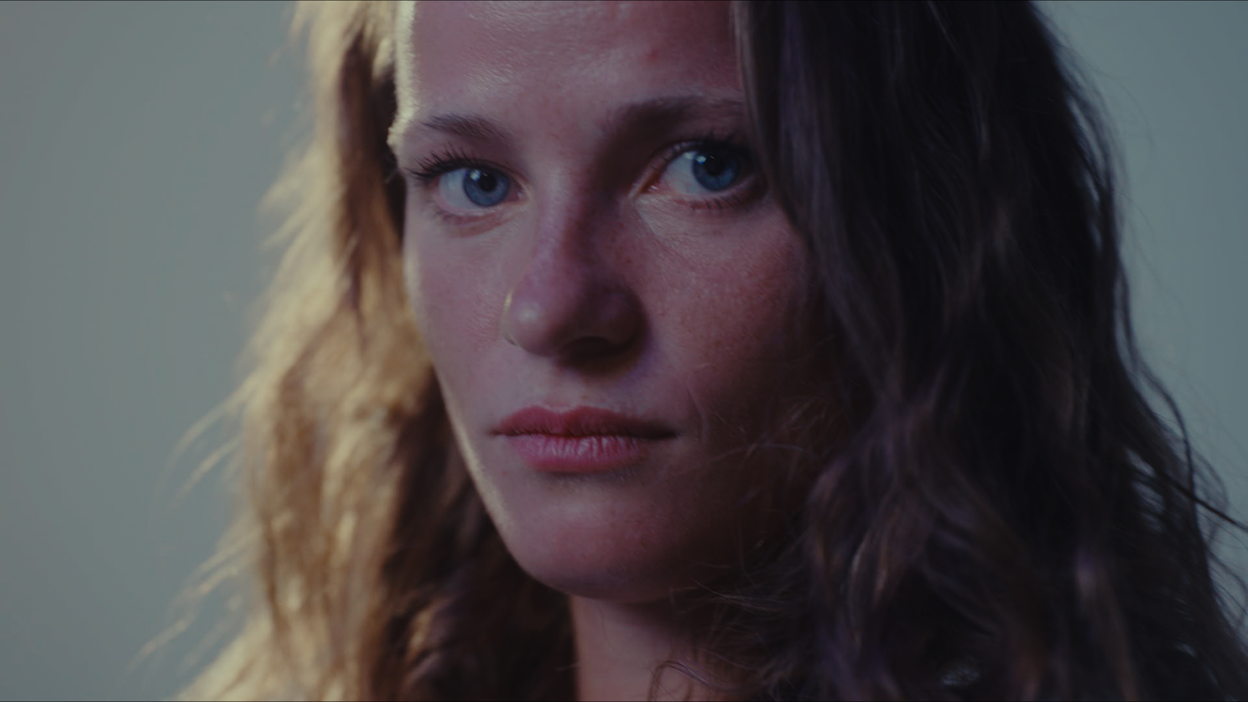 A close-up portrait of a young woman with blue eyes, fair skin, and wavy brown hair, looking directly at the camera.