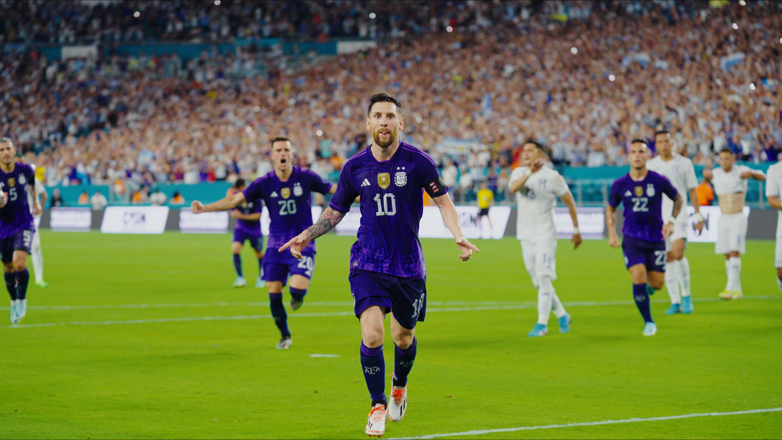 Soccer players celebrating on a field during a match, with a large crowd in the stadium background. The player in the foreground wearing a purple jersey with the number 10 is running towards the camera with a confident expression, while teammates and opponents celebrate behind him.