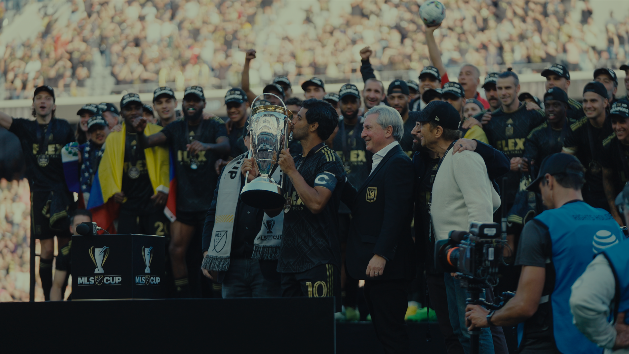 Soccer team celebrating victory on the stage after winning MLS Cup, with one player kissing the trophy, surrounded by teammates, team officials, and a camera crew in a stadium filled with spectators.