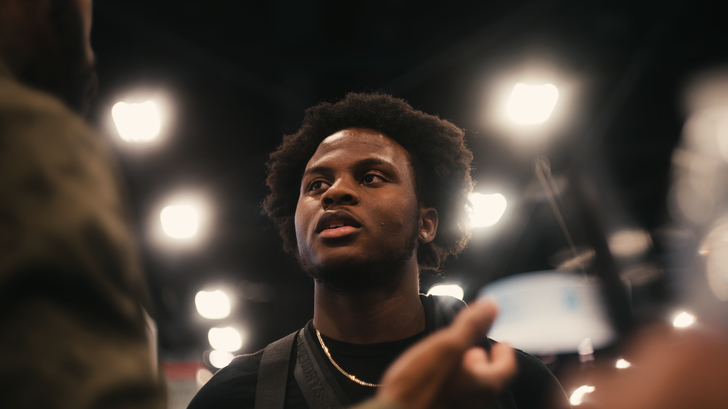A young man with curly hair and earrings looking to his right in an indoor setting with bright lights overhead.