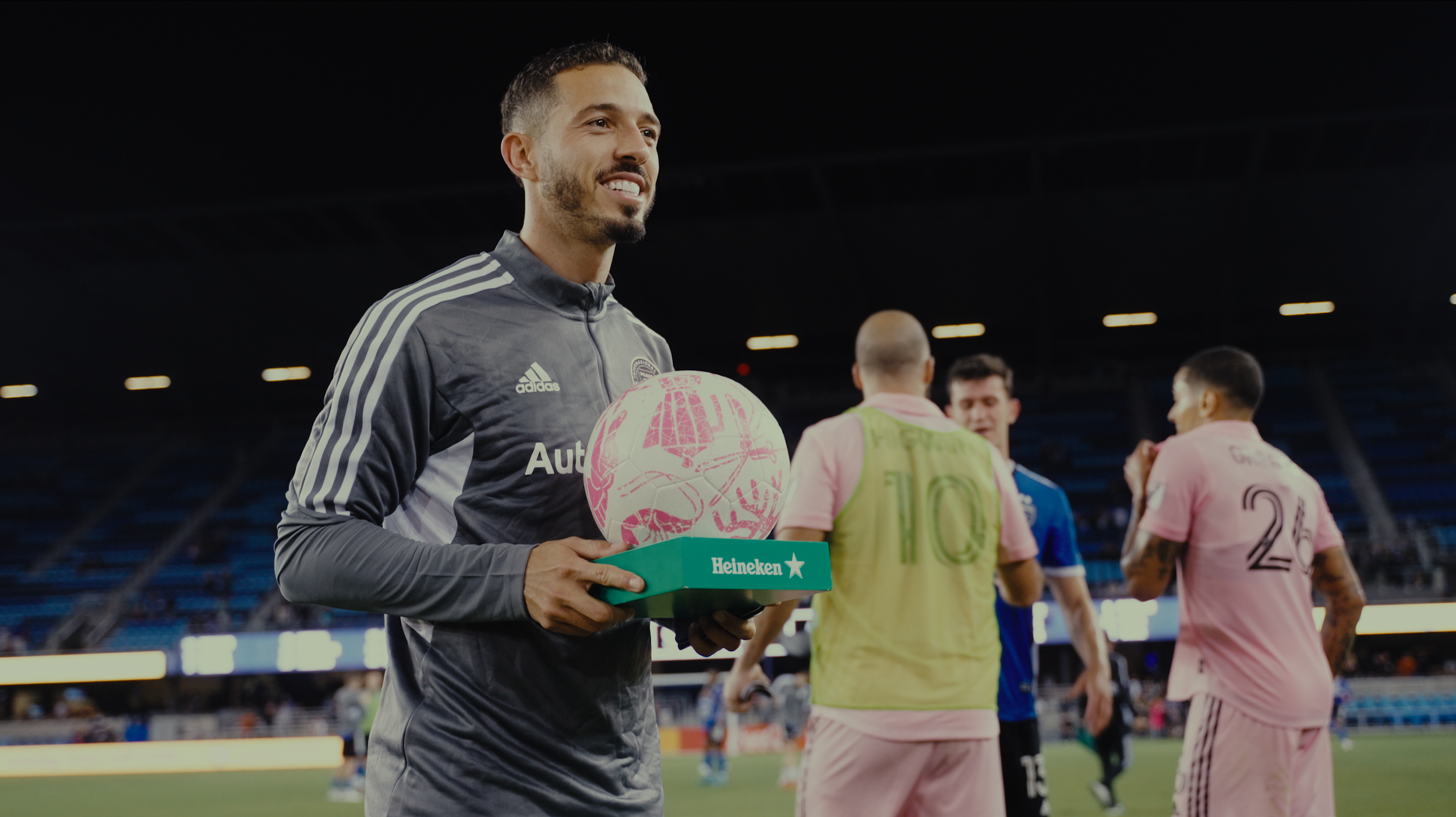 A male soccer player in a gray training jacket holding a pink and white soccer ball with Heineken branding and a green base, smiling after a game, with other players in pink and blue jerseys in the background in a stadium.