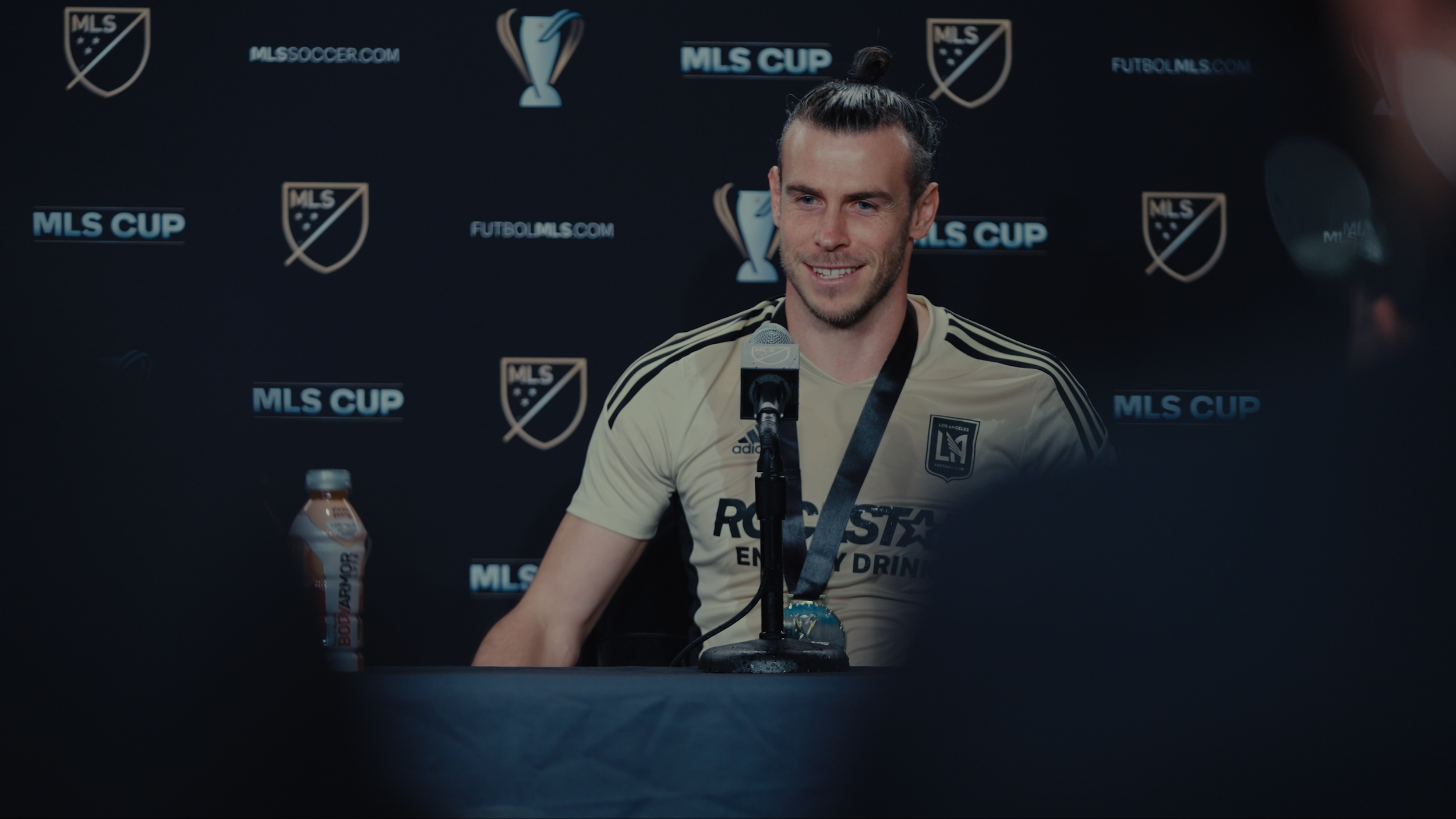 A man in a Los Angeles soccer jersey is smiling and speaking into a microphone during a press conference, with a backdrop displaying the MLS Cup logo and related branding.