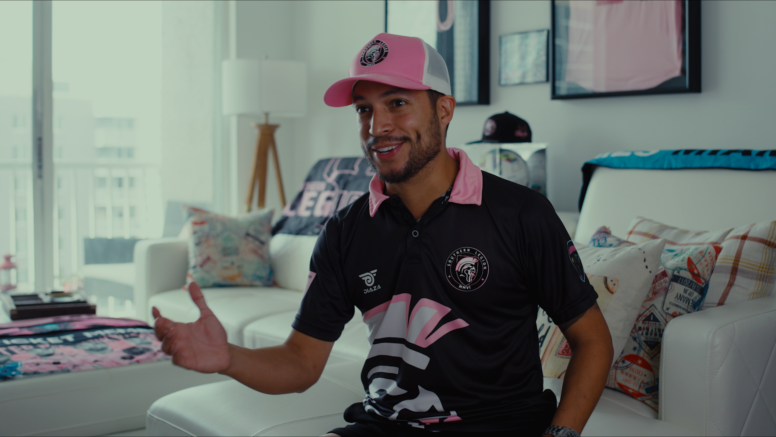 A man in a black and pink sports shirt and pink cap sitting on a white sofa in a bright living room, smiling and gesturing with his left hand.