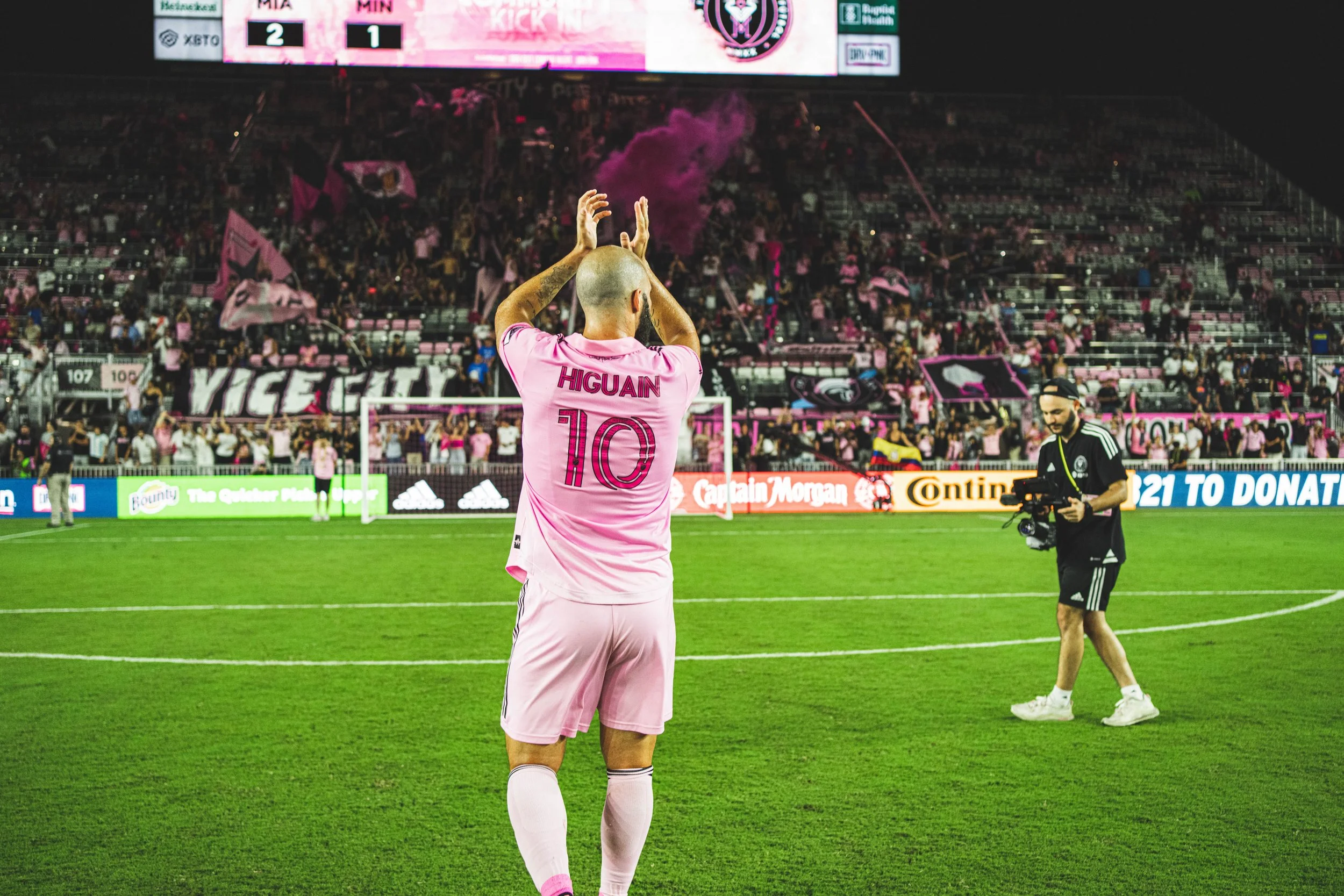 A soccer player wearing a pink uniform with the name 'HIGUAIN' and number 10 on the back is standing on a green field, facing away from the camera, with their hands raised above their head. The player is on a stadium field with fans and flags in the background, along with advertisements along the perimeter of the field.