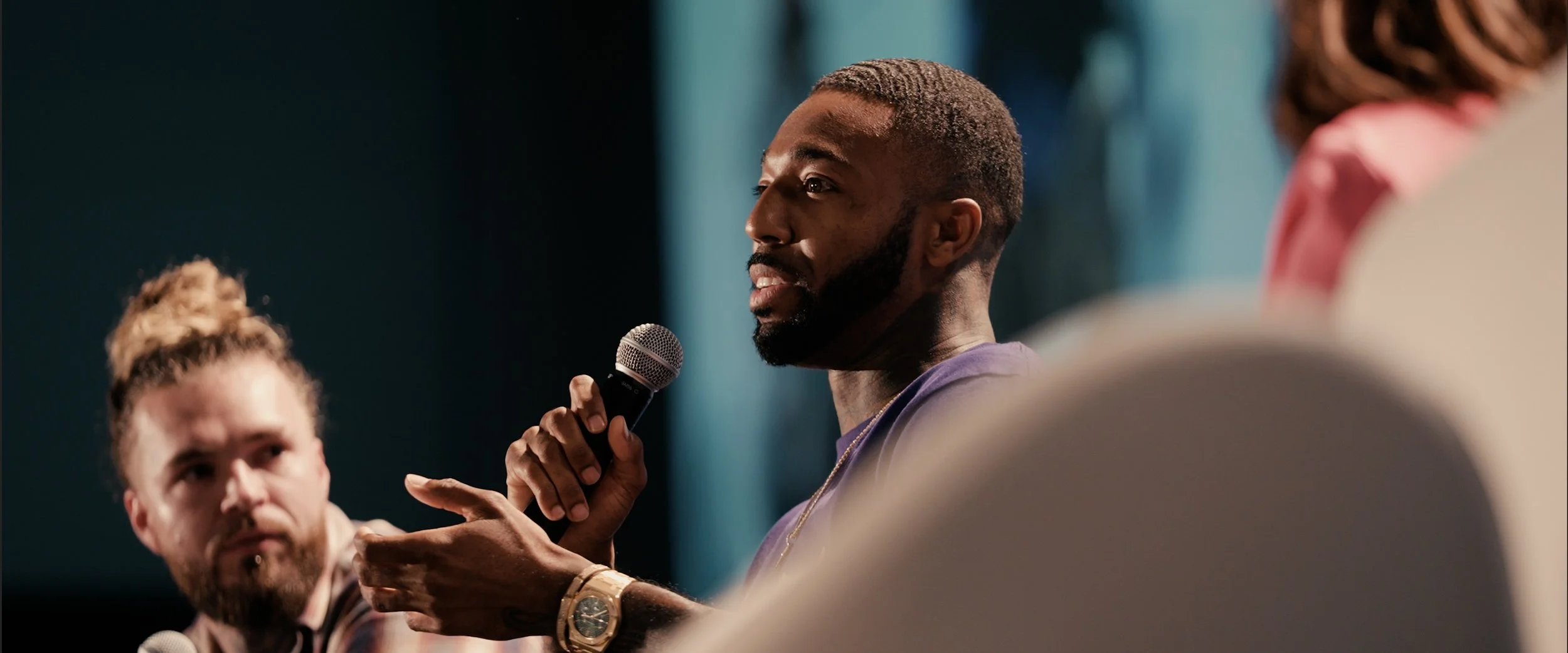 A man with a beard holding a microphone speaking during a panel discussion or conference, with a woman with light skin and blonde curly hair also visible, and additional blurred individuals in the background.