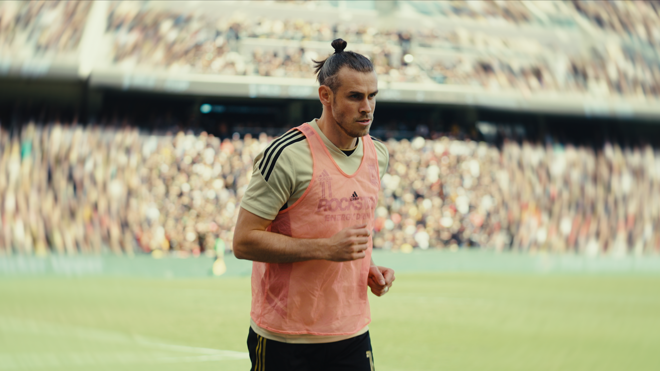 A male soccer player with a man bun, wearing a yellow jersey and pink training vest, on a soccer field during a game.