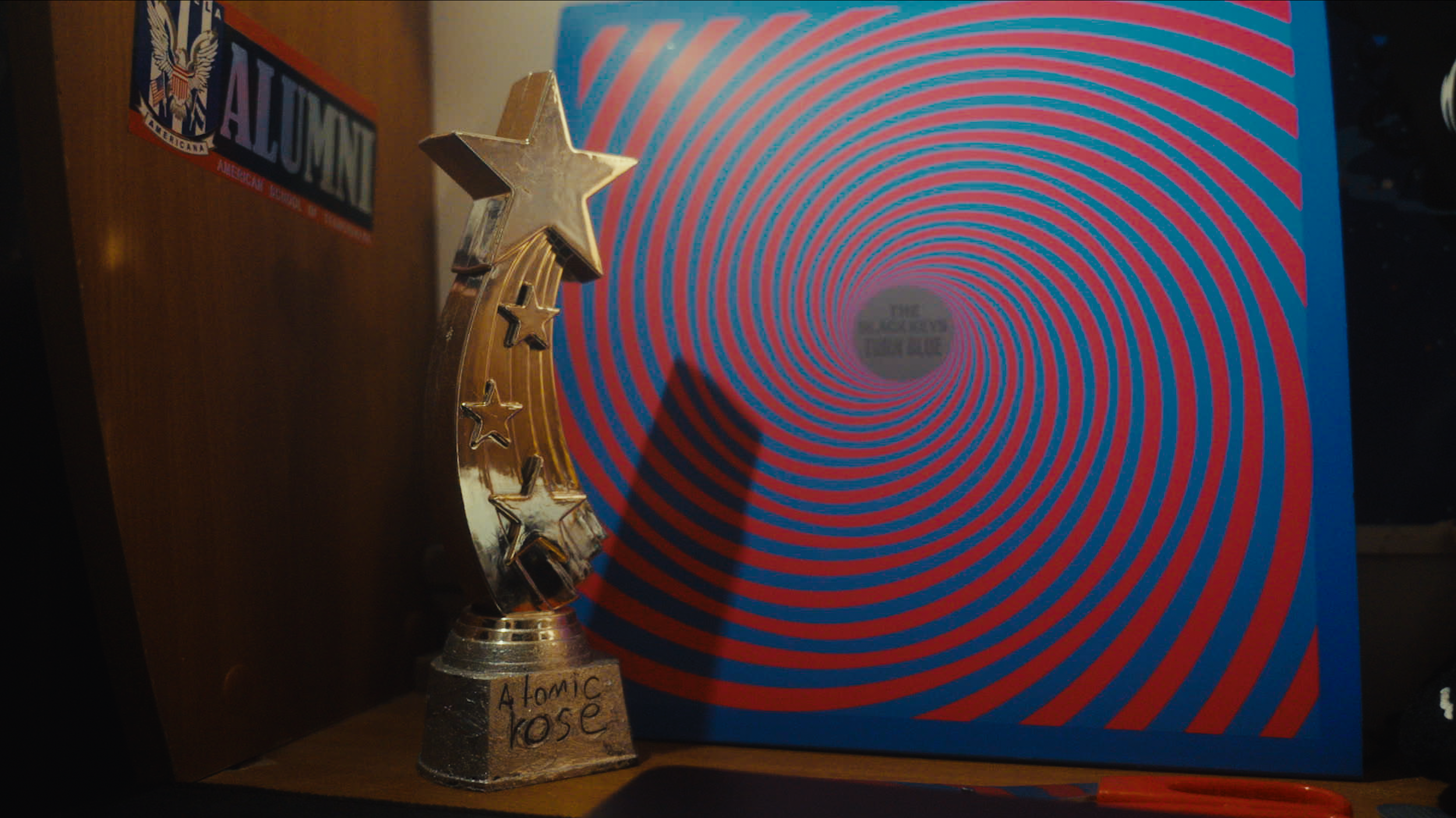 A gold trophy with stars and a sign that says "Aromatic Rose" in front of a blue and red hypnotic spiral background, next to a wooden plaque with an American flag sticker that reads "Alumni".