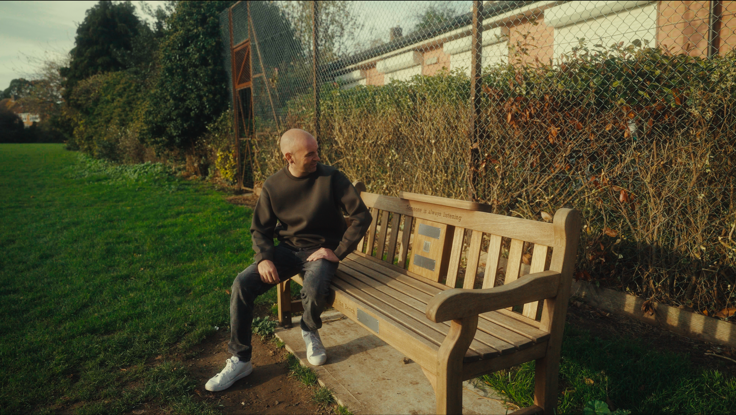 A bald man sitting on a wooden bench in a park, looking to his left, with a chain-link fence and brown bushes behind him. The bench has the phrase 'Someone is always listening' engraved on it.