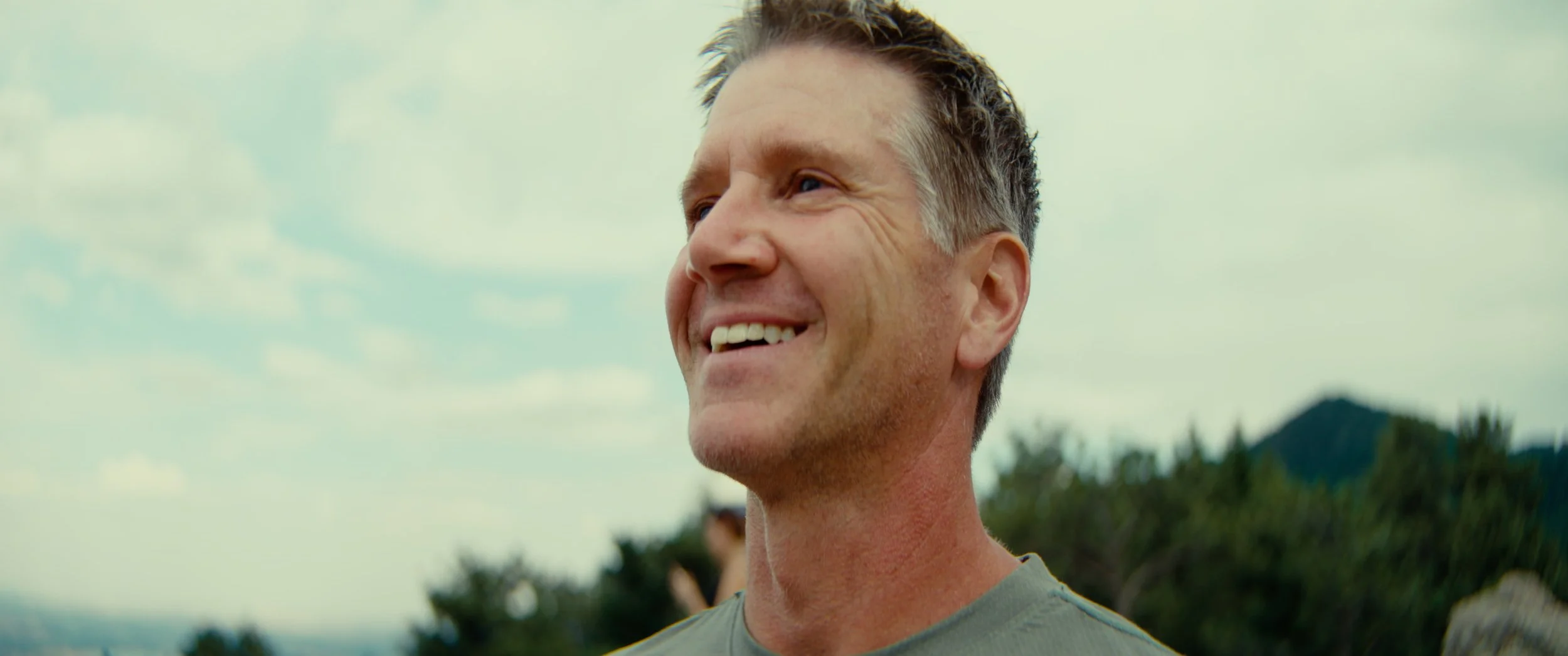 A middle-aged man with short, dark hair smiling outdoors on a cloudy day, with trees and a distant mountain in the background.
