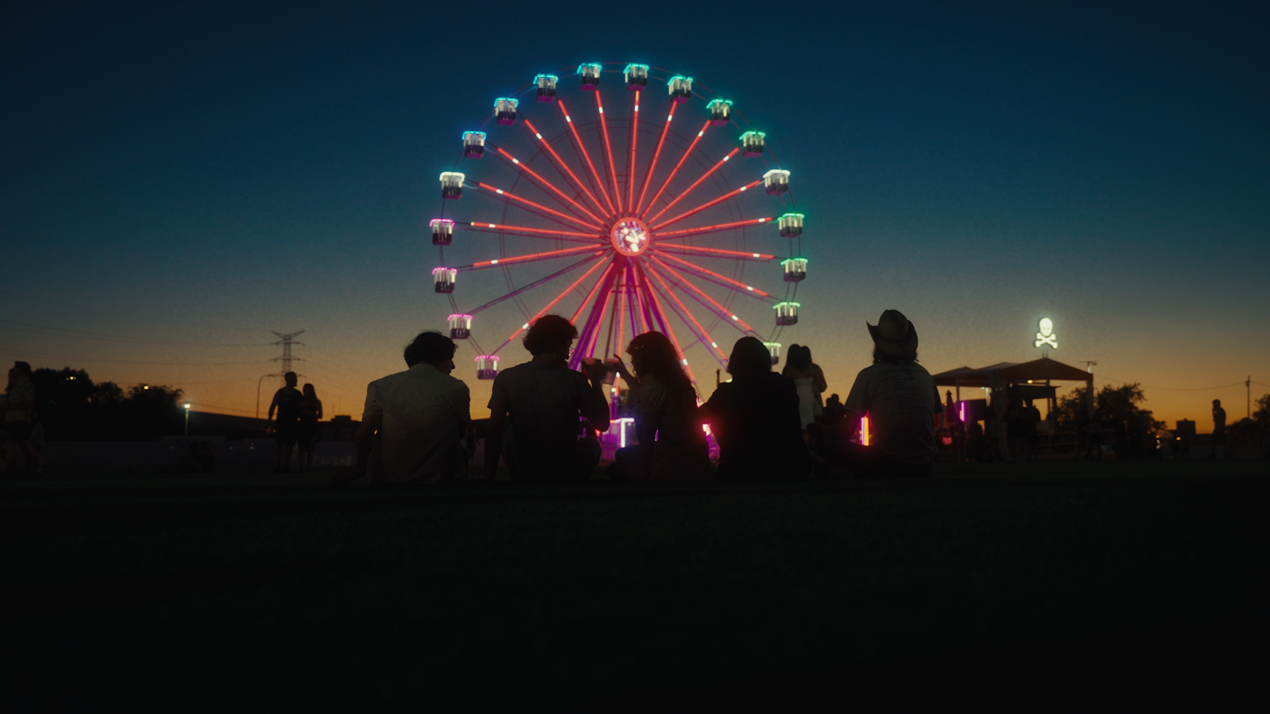 People sitting on the ground near a lit-up Ferris wheel at dusk, with a sunset sky in the background and a ghost skull figure on a structure nearby.