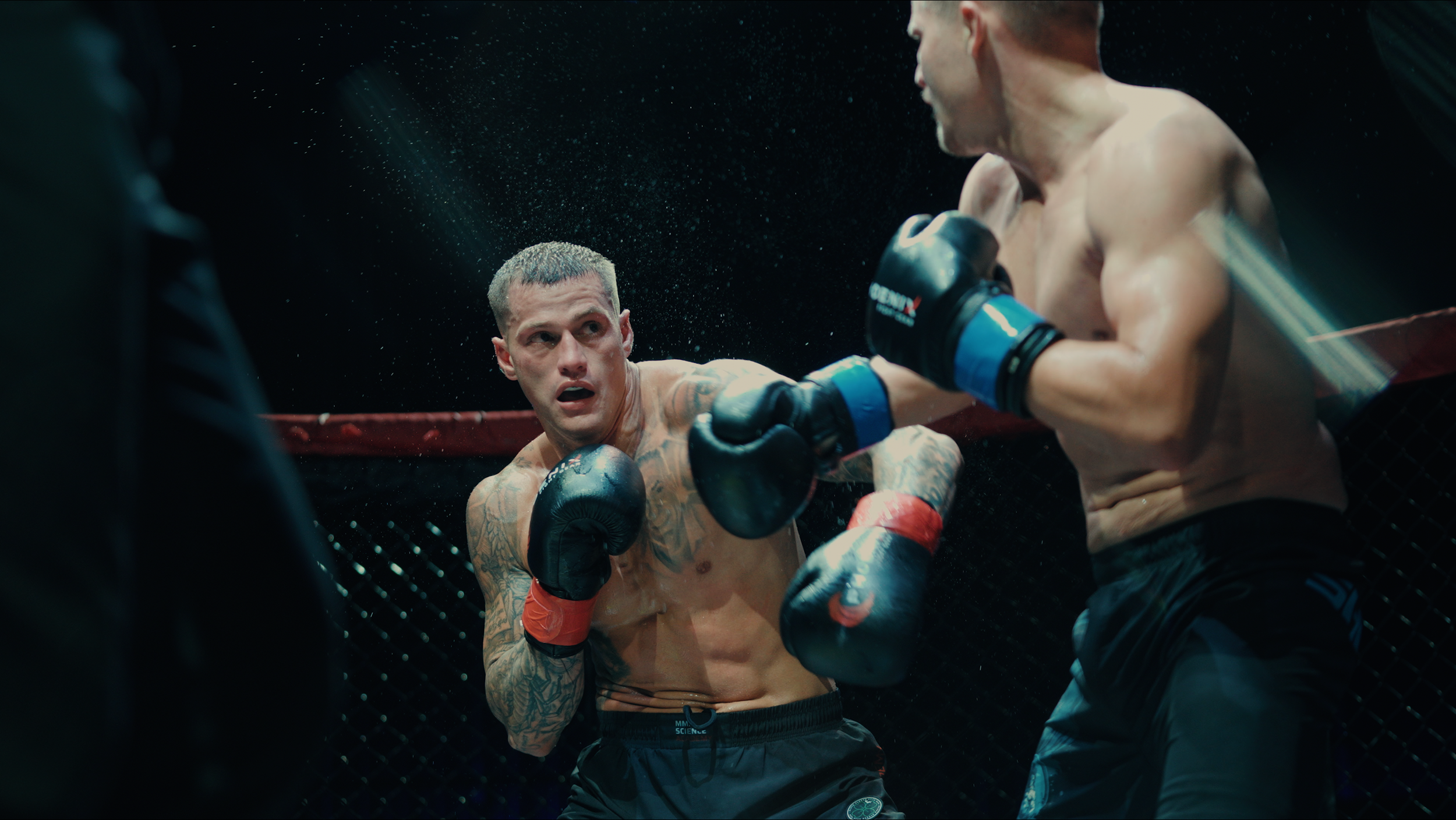 Two male mixed martial arts fighters inside a cage engaged in a fight, with one fighter throwing a punch. The fighter on the left has tattoos and a focused expression, while the fighter on the right is blocking the punch. The background is dark and the fighters are illuminated by overhead lighting.