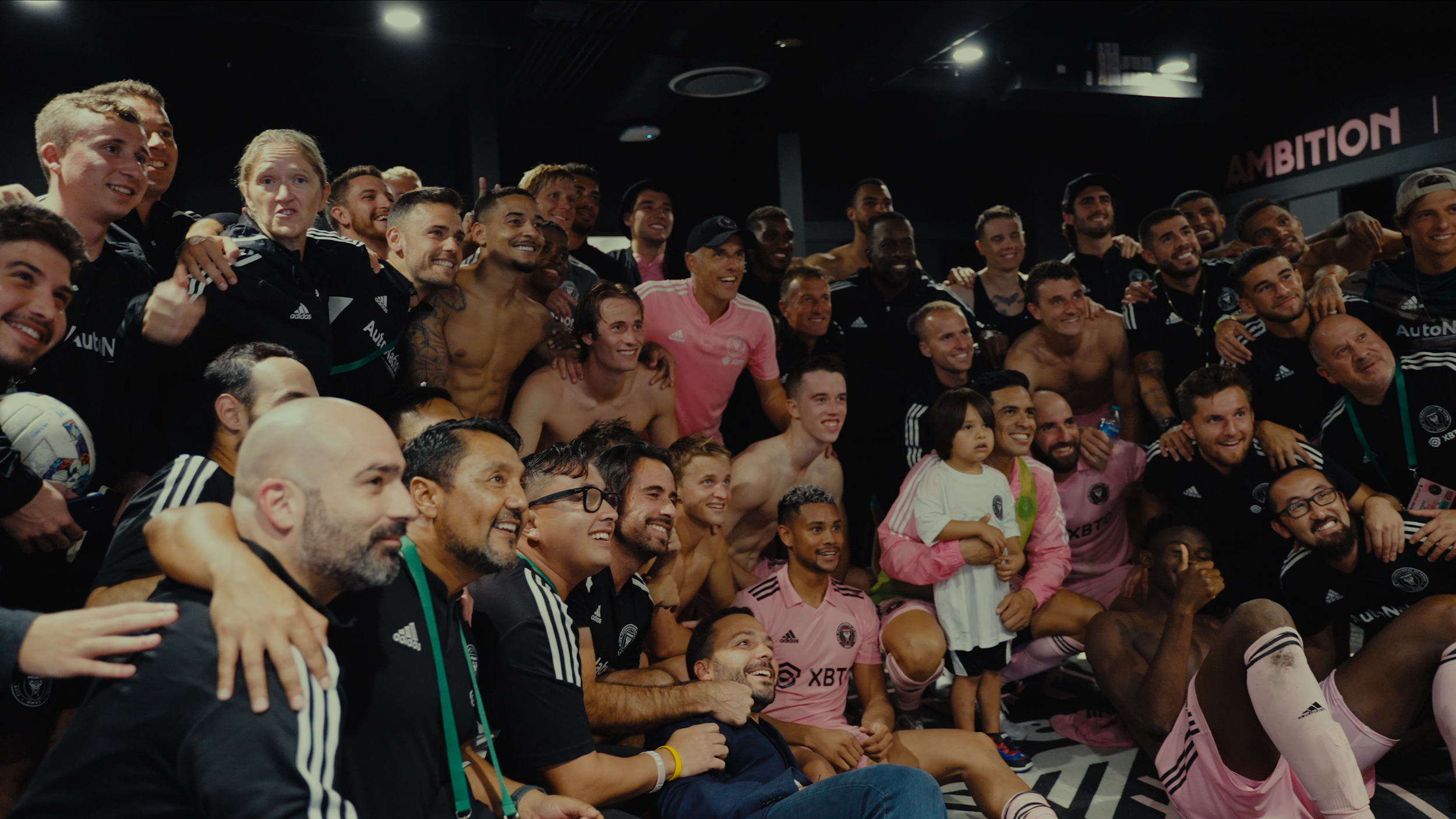 Group of soccer players and staff posing for a photo inside a team room, some are shirtless, wearing black or pink jerseys with a mix of seated and standing positions, celebrating and smiling.