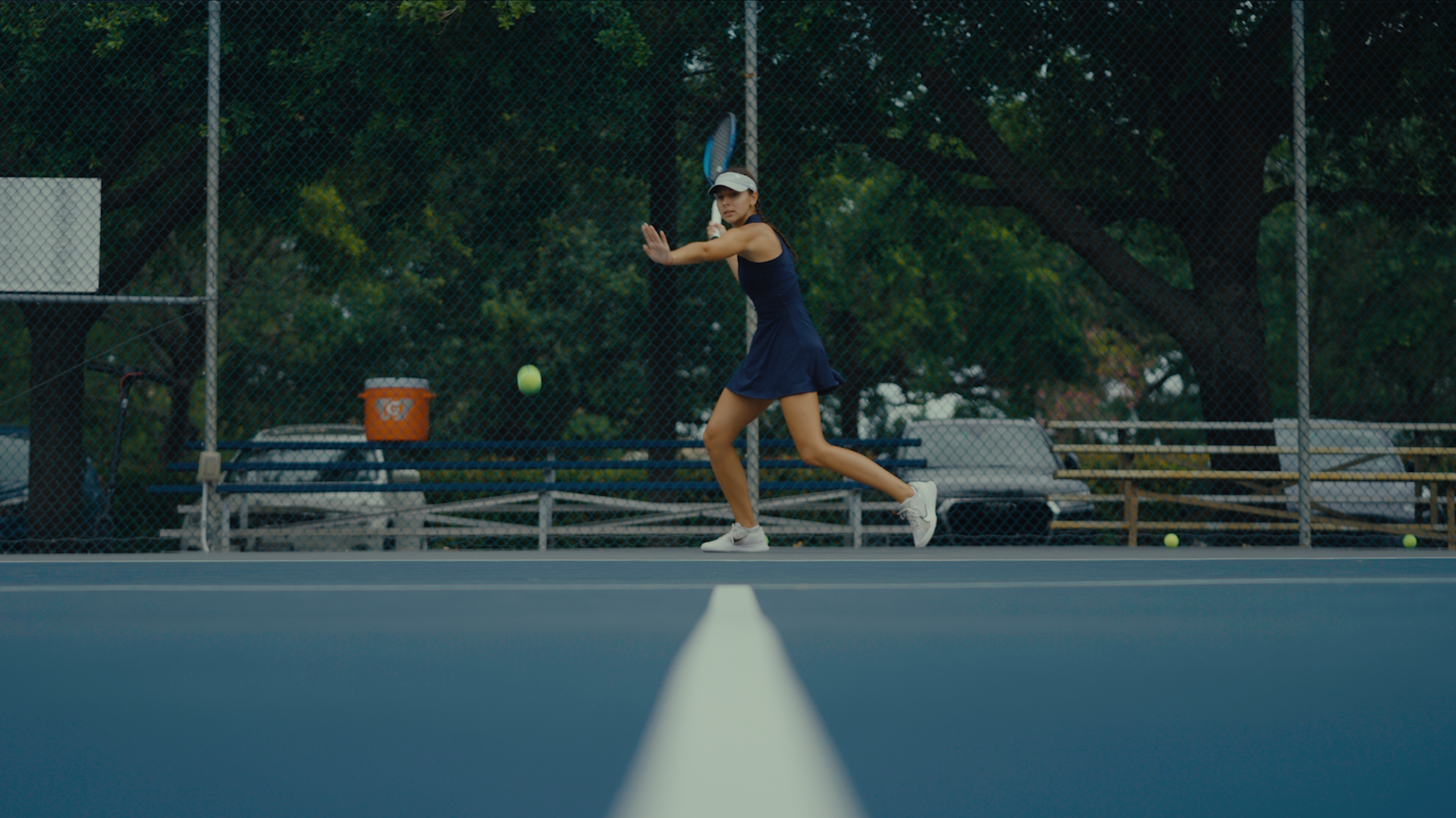 Tennis player in a blue dress preparing to hit a tennis ball on a court surrounded by a fence.