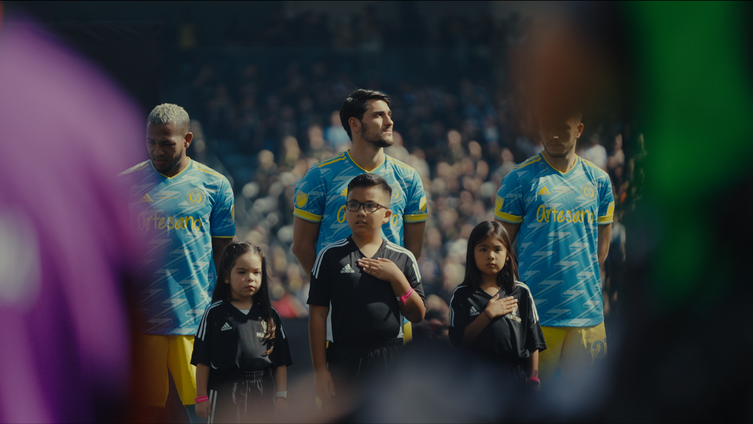 Soccer players and children standing during national anthem at a stadium, with spectators in the background.