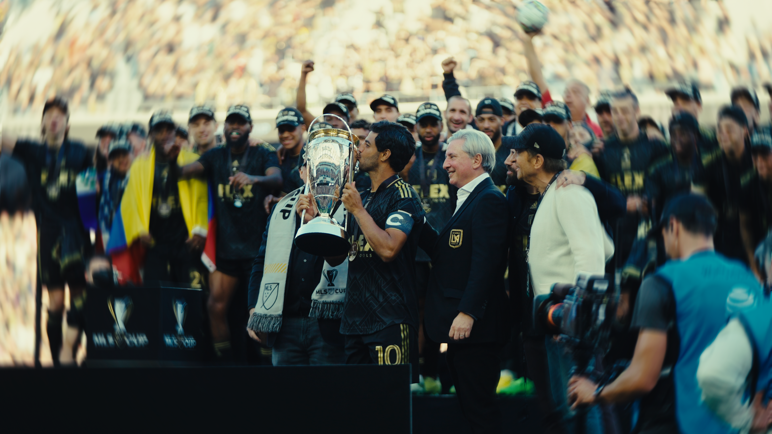 Soccer team celebrating victory with trophy on field, surrounded by fans with cascade of cheering supporters in background.