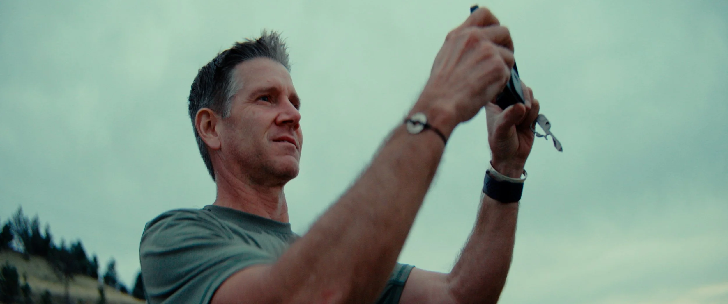 A man with short gray hair wearing a gray t-shirt and a black bracelet on his right wrist, holding and looking at a small camera or phone outdoors, with a cloudy sky in the background.