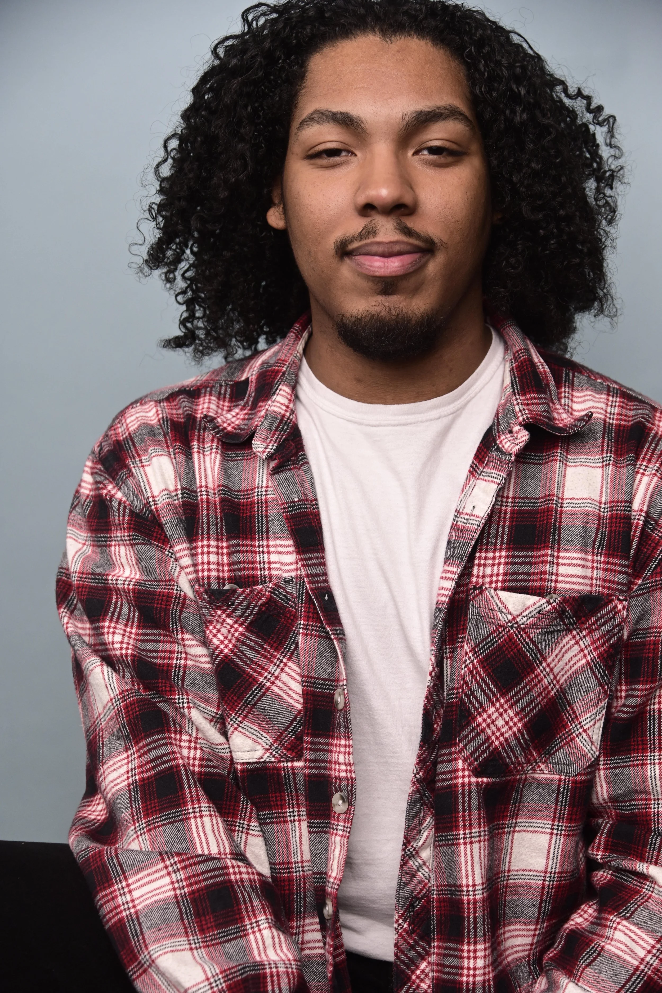 A young man with medium skin tone, curly black hair, and a small goatee, smiling slightly, wearing a red and black plaid shirt over a white t-shirt, standing against a light blue background.