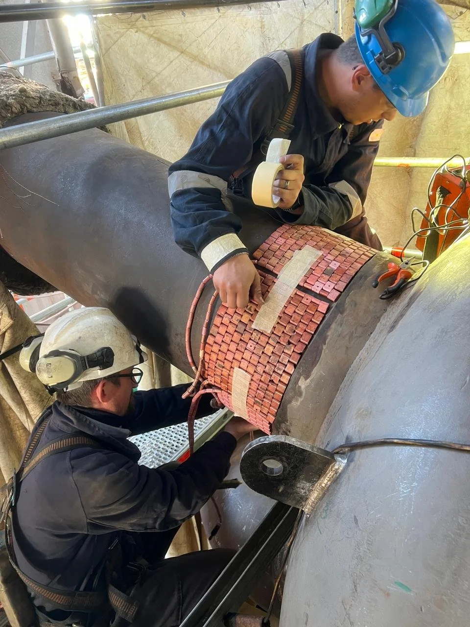 Dos trabajadores usando equipo de protección, inspeccionando y trabajando en una tubería grande con aislante térmico, en un entorno industrial.