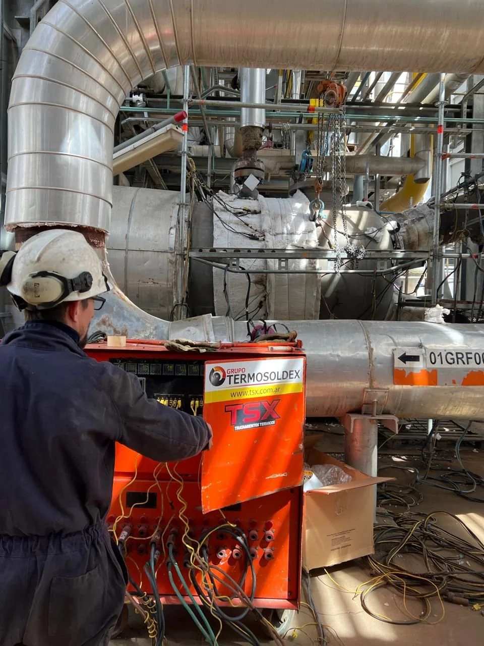 Trabajador con casco revisando un sistema de control eléctrico en una planta industrial con tuberías de metal y maquinaria.