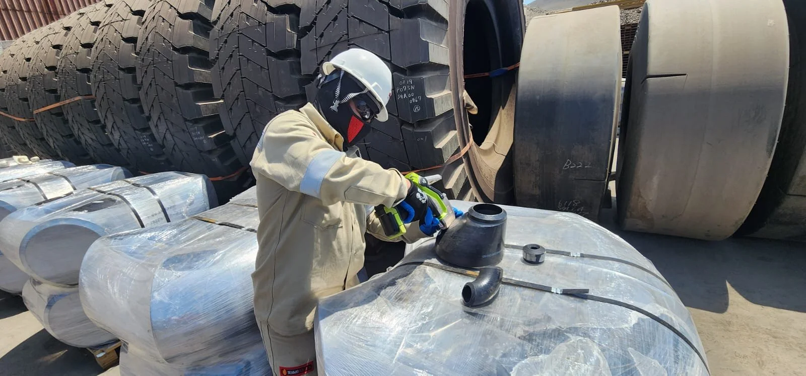 Trabajador usando equipo de protección, inspeccionando piezas de maquinaria en un área de almacenamiento de neumáticos y componentes industriales.