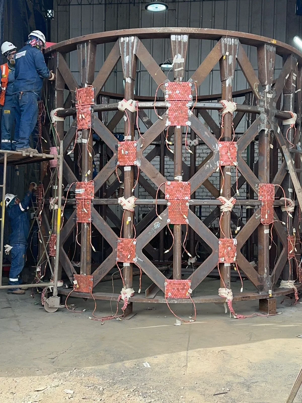 Estructura metálica de gran tamaño en proceso de ensamblaje, con varios trabajadores en un taller, usando equipo de protección y soldando partes de la estructura.