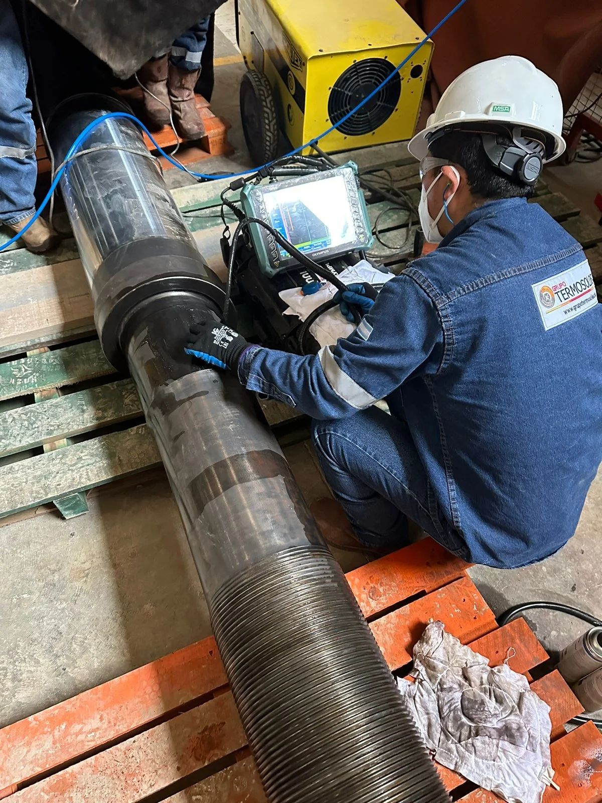 Técnico con casco, gafas y máscara revisando un tubo grande con equipo de medición portátil en una planta industrial.