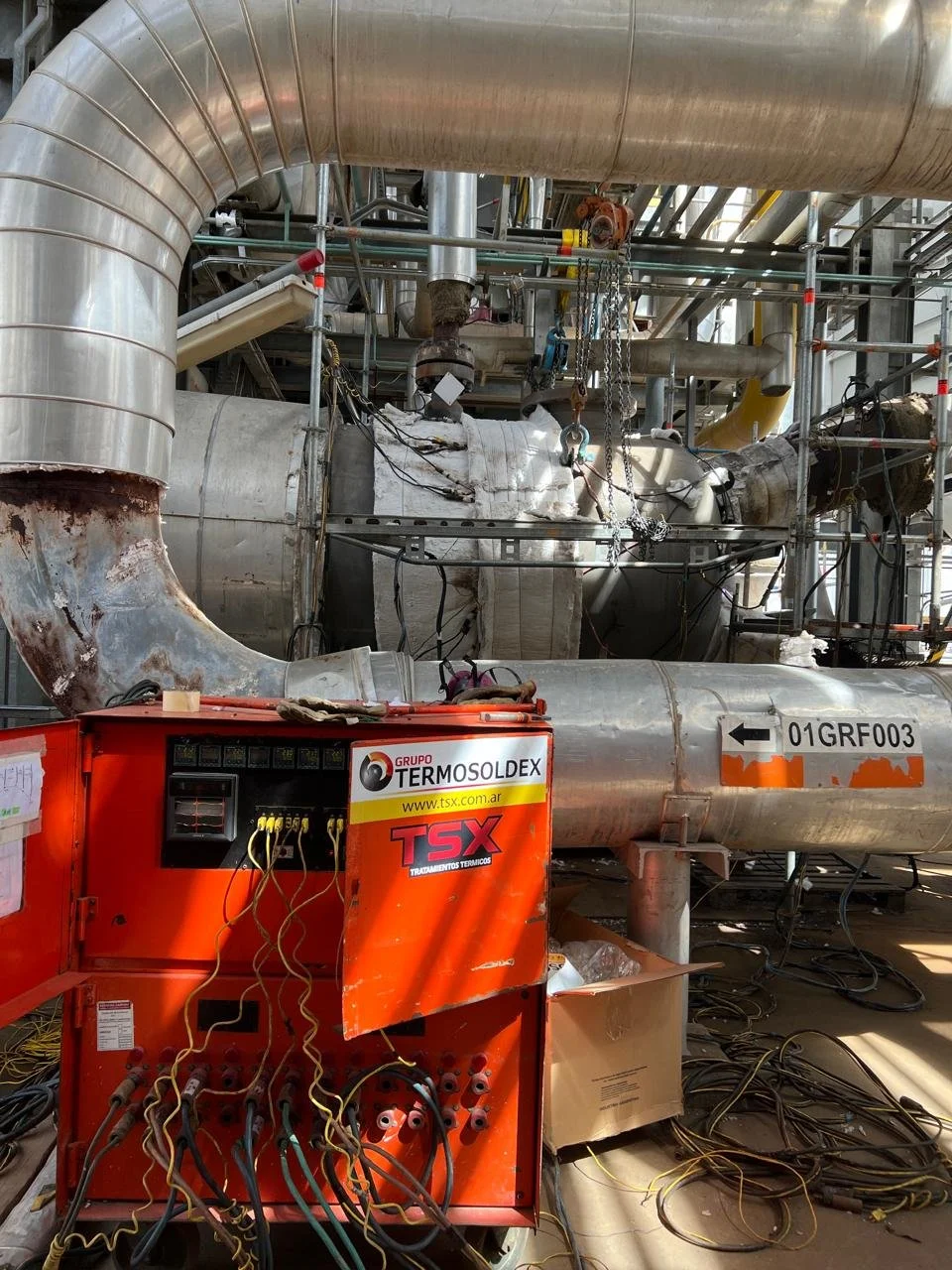 Instalación industrial con tuberías metálicas, maquinaria y cables eléctricos, en un entorno de planta de procesamiento o fabricación.