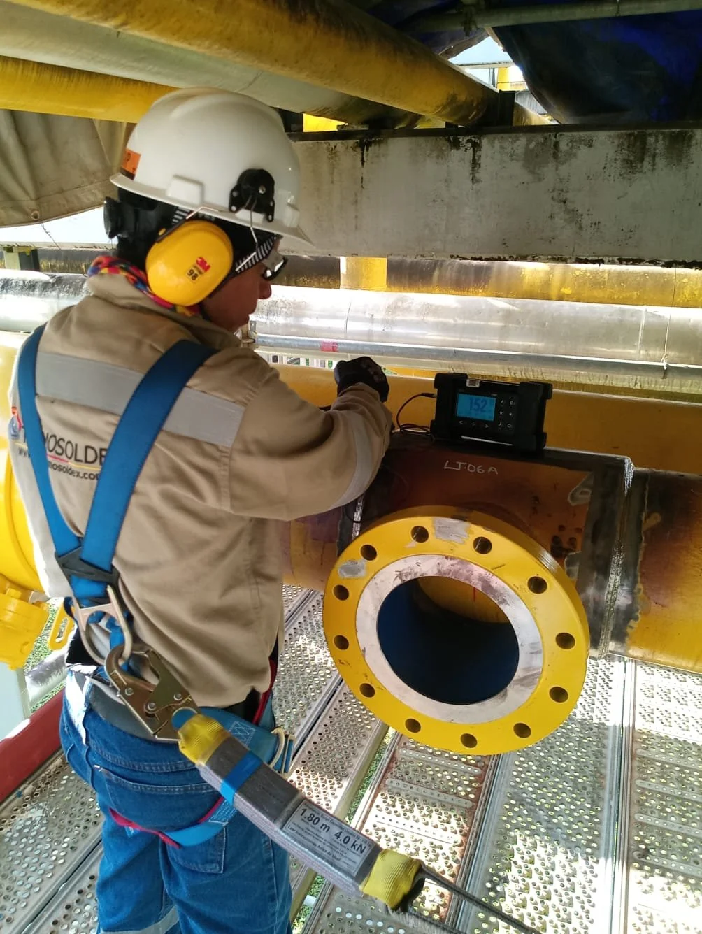 Un trabajador con casco y protección auditiva inspecciona una tubería grande en una estructura de acero en una obra de construcción.