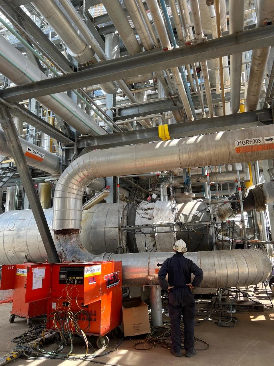 Un trabajador en casco y ropa de protección inspecciona una gran estructura de tuberías metálicas industriales en una planta de ingeniería.