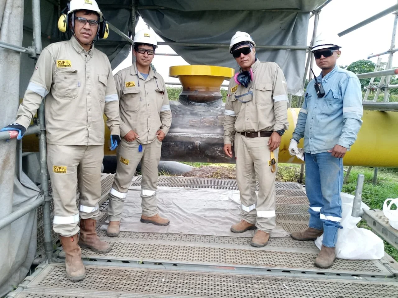 Cuatro ingenieros o técnicos en ropa de protección y cascos, de pie frente a una gran tubería en una plataforma de estructura metálica en un entorno de trabajo. Están usando gafas de protección y guantes.