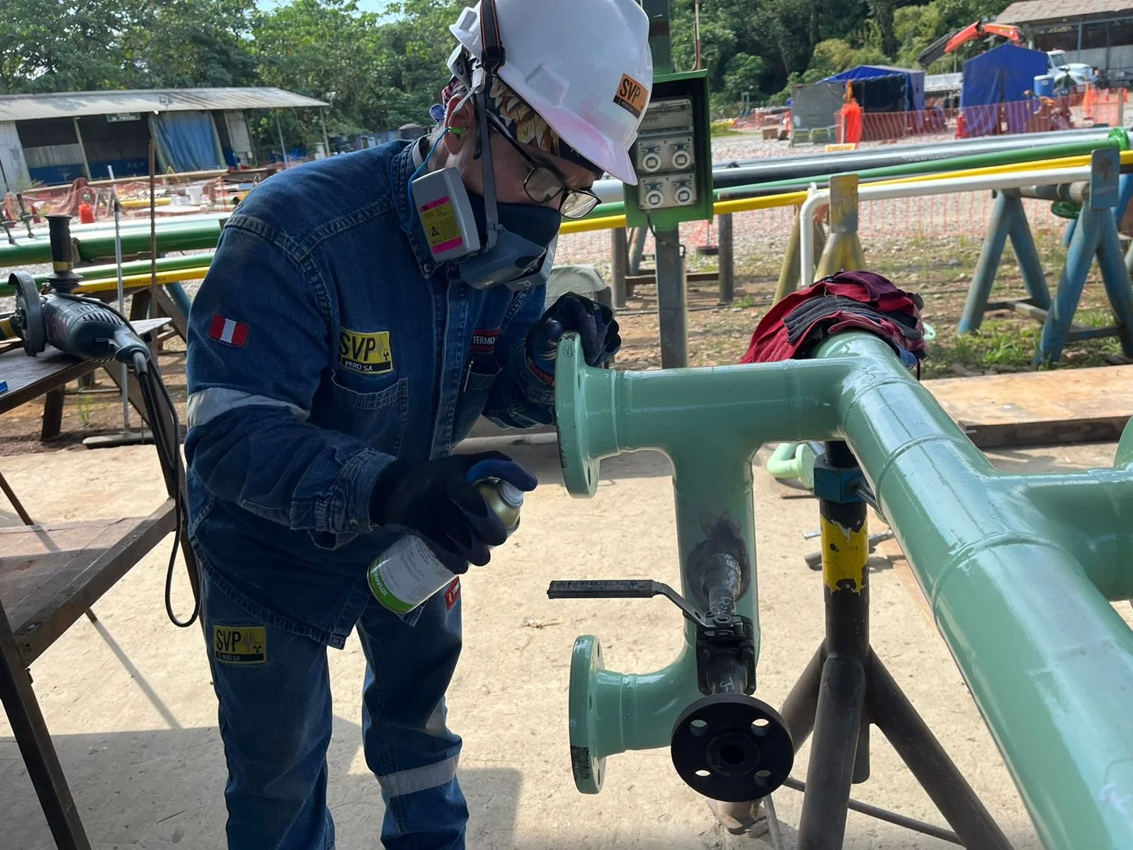Un trabajador con equipo de protección plásticos, casco y gafas, lacando una tubería de color verde afuera en un lugar de construcción.