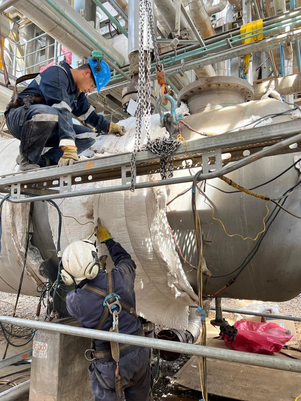 Dos técnicos trabajan en la reparación de una tubería grande en una planta industrial, con mucho equipo y tuberías alrededor.
