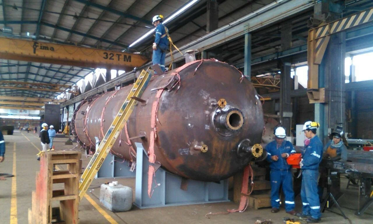 Personal trabajando en la construcción o reparación de un gran recipiente de metal en una fábrica, con equipo de protección y un grúa de alta capacidad.